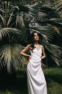 Elegant sensual woman brunette bride in a silk wedding dress posing among palm trees in the park