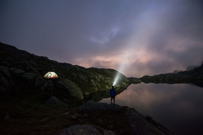 Rear view of man with flashlight standing on rock against sky