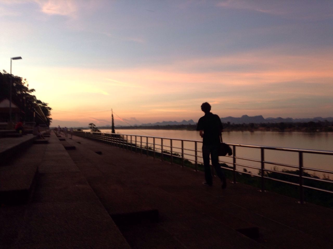 sunset, sky, silhouette, water, railing, men, full length, rear view, built structure, lifestyles, architecture, standing, transportation, cloud - sky, walking, building exterior, bridge - man made structure, orange color