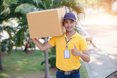 Portrait of smiling delivery person with box showing thumbs up on footpath