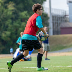 Full length of boy playing goalkeeper in soccer game on grass with coach and teammate in background