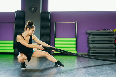 Full length of woman holding ropes while crouching on floor in gym