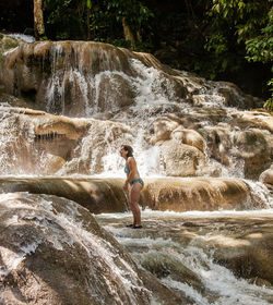 Side view of woman wearing bikini standing on rock against waterfall