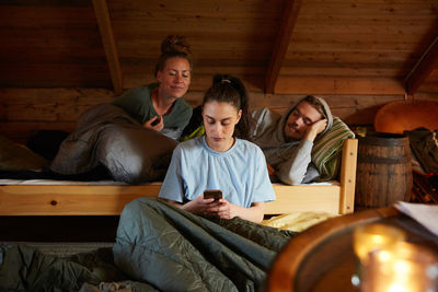 Friends looking at female using smart phone while relaxing on bed in cottage