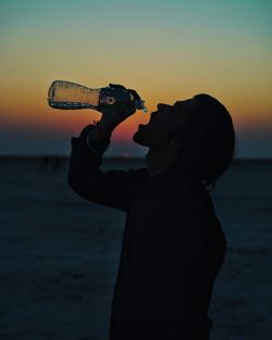 Side view of man holding camera at beach against sky during sunset