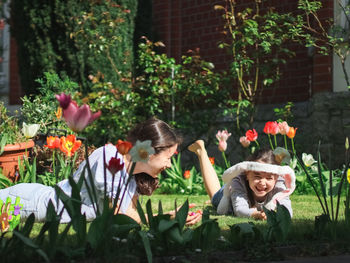 Two beautiful caucasian girl sisters with happy emotions lie on the lawn with flowers in the yard.