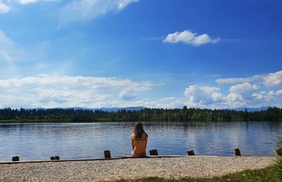 Rear view of woman sitting by lake against sky