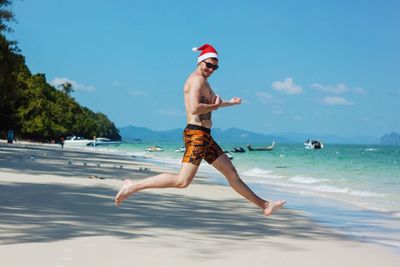 Shirtless man wearing santa hat while jumping on sand against sea at beach