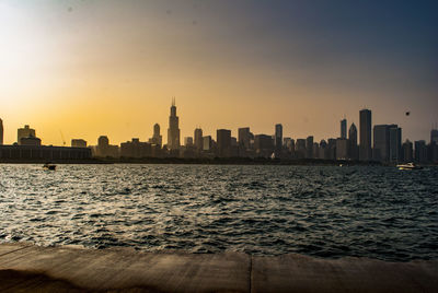 Sea by cityscape against sky during sunset