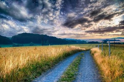 Road passing through field against cloudy sky