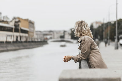 A middle-aged blonde woman with curly hair stands on the river embankment in the city.