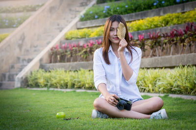 Full length of woman holding leaf and camera while sitting on grassy field at park