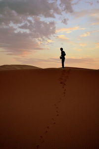 Silhouette young man standing on desert against cloudy sky during sunset