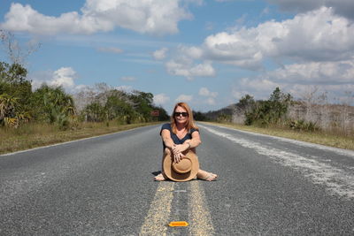 Portrait of woman sitting on empty road against sky