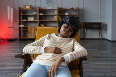 Black young woman removed virtual reality goggles and fell asleep after watching series in armchair.