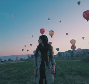 Young woman standing on field with hot air balloons flying against clear sky during sunset