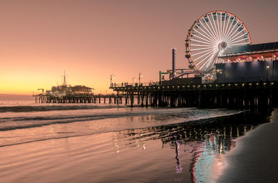 Illuminated ferris wheel by sea against sky at sunset