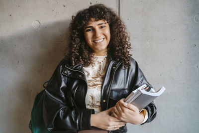 Portrait of smiling young woman holding book and backpack leaning on gray wall