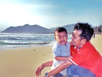 Father looking at crying son on beach against sky