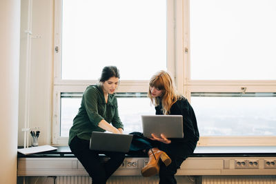 Confident female colleagues discussing over laptops while sitting on window sill at creative office