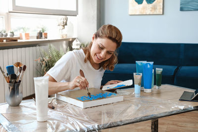 Woman artist painting picture at home with acrylic paints and spatula over the window. adult hobby