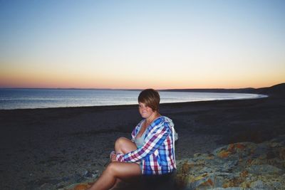Young woman sitting at beach against clear sky during sunset