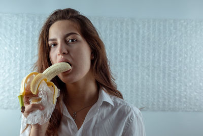Portrait of young woman eating banana against wall