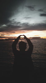 Silhouette young man making heart shape while standing at beach against cloudy sky during sunset