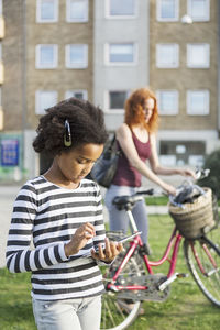 Girl using mobile phone with mother holding bicycle in background