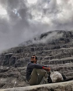 Low angle view of young man sitting on mountain against cloudy sky