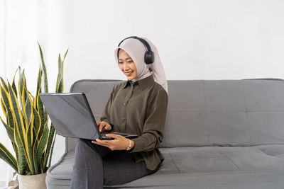 Portrait of young woman using laptop while standing against wall