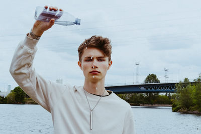 Portrait of confident young man pouring water from bottle against sky