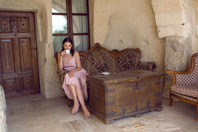 Woman sits on an old sofa at the entrance to the house in the rock and drinks tea