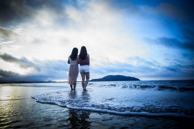 Rear view of friends standing at beach against cloudy sky during sunset