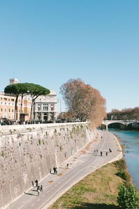 People walking on road by river in city against clear blue sky