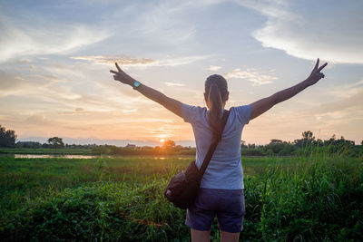 Woman with arms outstretched standing on field against sky during sunset