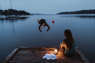 Man diving in water while woman sitting on pier by sea against sky