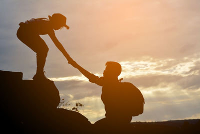 Silhouette boy assisting girl while hiking against sky during sunset