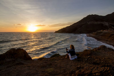 People sitting on rock by sea against sky during sunset