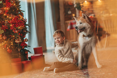 Candid authentic happy little boy in knitted beige sweater hugs dog with bow tie at home on xmas