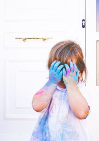 Girl with colored powder covering face with hands standing against door