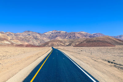 Road leading towards mountains against clear blue sky