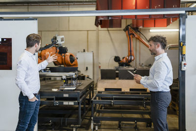 Businessman having discussion with colleague while standing by robotic arm machine at factory