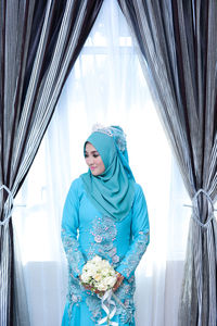 Bride holding flower bouquet while standing against window