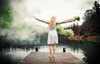 Rear view of woman holding distress flare standing by lake against sky