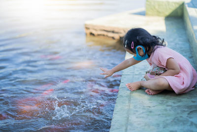 Cute girl wearing protective face mask feeding carp koi fishes at pond.