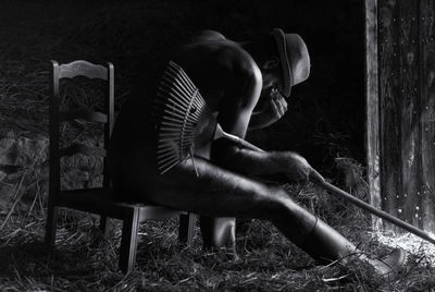 Naked sad man sitting on chair with rake in barn