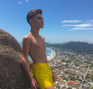 Shirtless young man standing by rock with cityscape in background against blue sky