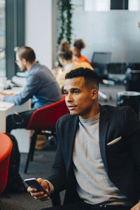 Businessman holding mobile phone while looking away against colleagues sitting at office