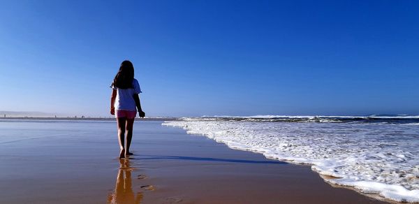 Rear view of woman standing on beach against clear blue sky